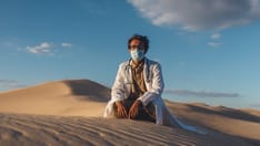 photo of A doctor in a white coat, mask, and stethoscope sits alone in a vast sand desert under a clear blue sky.
