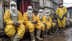 photo of Health workers in yellow hazmat suits and masks sit together in a muddy alleyway next to dilapidated buildings