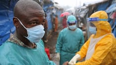 photo of Healthcare workers in protective gear, including gowns and face shields, assist people outdoors during the rain.