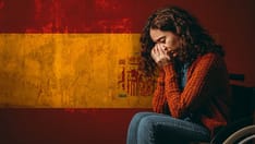 photo of A young woman sits in a wheelchair, crying into her hands against a backdrop of a weathered Spanish flag.
