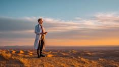 photo of A doctor in a white coat stands on a high desert dune, looking out over a vast, sandy landscape at sunset.
