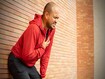 Sportsman wearing a red jacket leaning against a brick wall of clutching his chest acute pain possible heart attack. Hands male holding chest with symptom heart attack disease on city street.