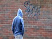 Photo showing a teenage boy / youth wearing a blue hoodie and standing beside a brick wall, in a run-down part of the city.