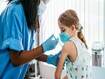 Young girl watching her being injected with COVID-19 vaccine at a medical clinic. African American female nurse or doctor injecting vaccine into caucasian blonde girl patient siiting on examination table in vaccination center.