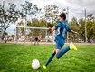 Rear view of agile 13 year old boy athlete wearing blue uniform and approaching ball for kick toward goal.